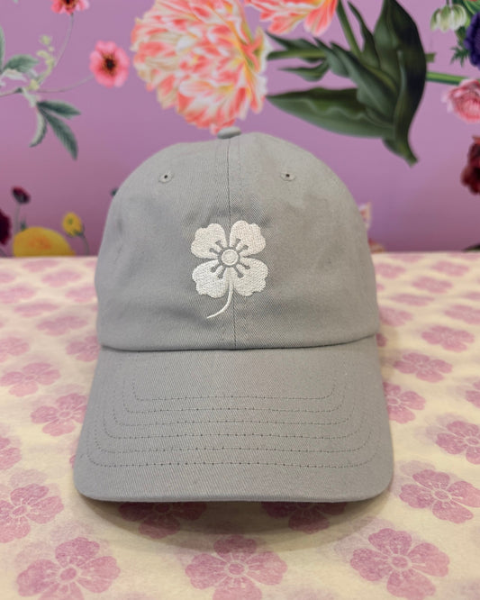 A light gray baseball cap with an embroidered white flower on the front, displayed on a pink patterned surface against a floral pink background.