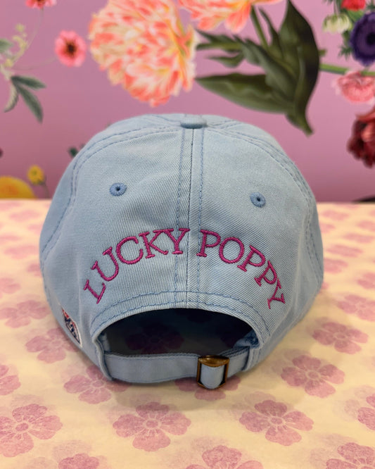 A light blue baseball cap with 'LUCKY POPPY' embroidered in dark pink on the side, displayed on a floral-patterned surface.