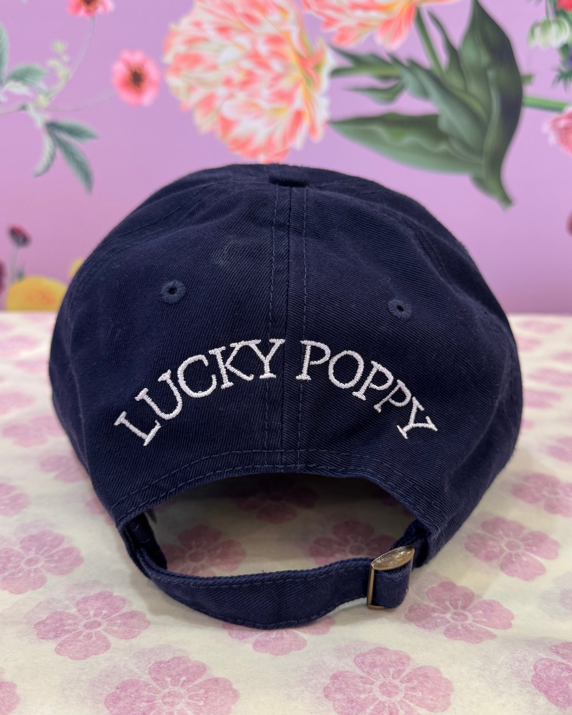 A navy blue baseball cap with 'LUCKY POPPY' embroidered in white on the side, displayed on a floral-patterned surface.