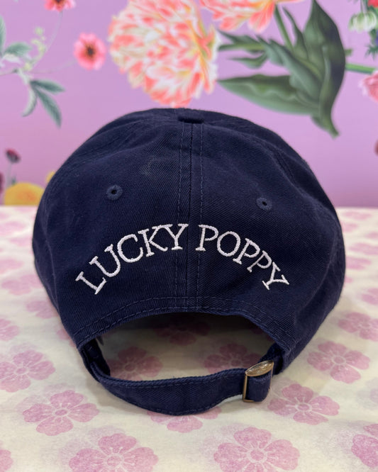 A navy blue baseball cap with 'LUCKY POPPY' embroidered in white on the side, displayed on a floral-patterned surface.