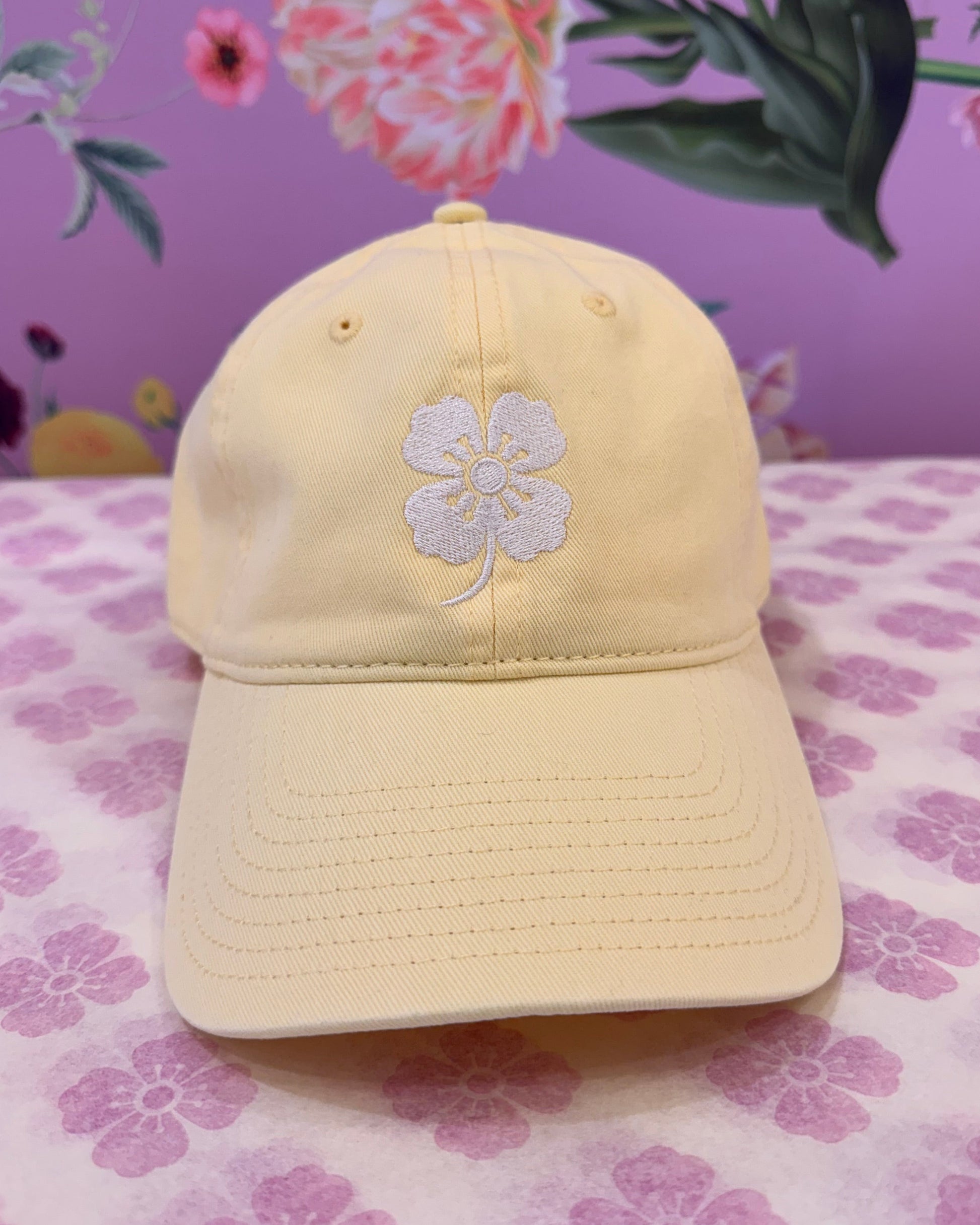 A pale yellow baseball cap with a white embroidered flower on the front, displayed on a pink and white floral patterned fabric next to a purple floral background.
