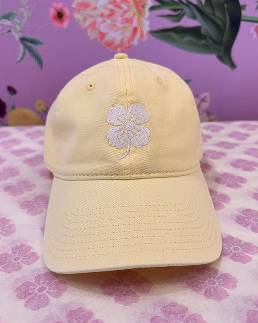 A pale yellow baseball cap with a white embroidered flower on the front, displayed on a pink and white floral patterned fabric next to a purple floral background.