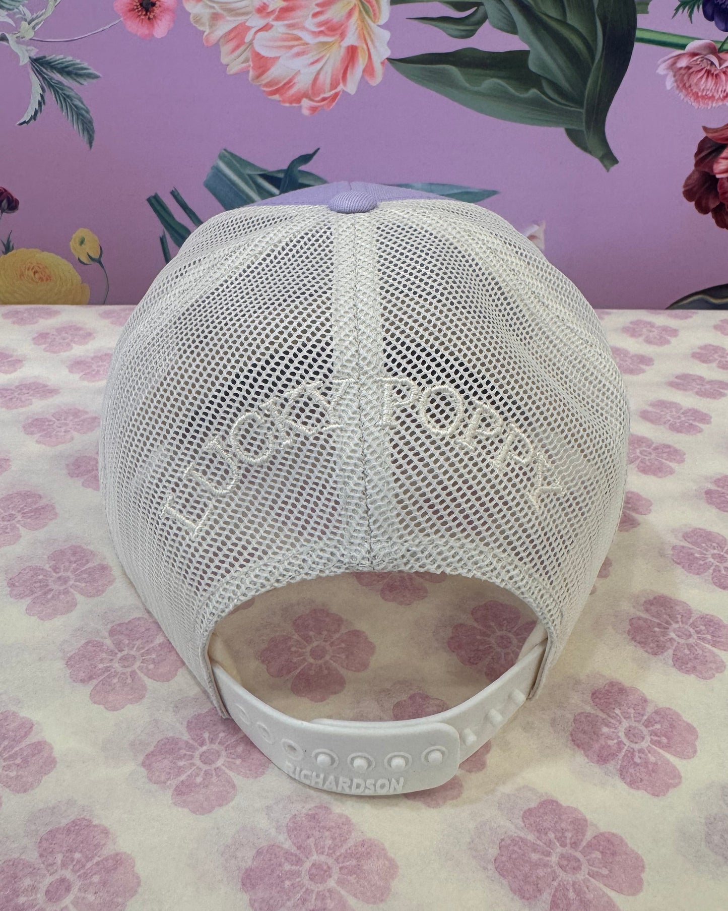 A white mesh baseball cap with 'LUCKY POPPY' printed on the front, resting on a split floral-patterned surface with a purple background and a cream-and-pink floral background.