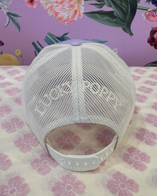 A white mesh baseball cap with 'LUCKY POPPY' printed on the front, resting on a split floral-patterned surface with a purple background and a cream-and-pink floral background.