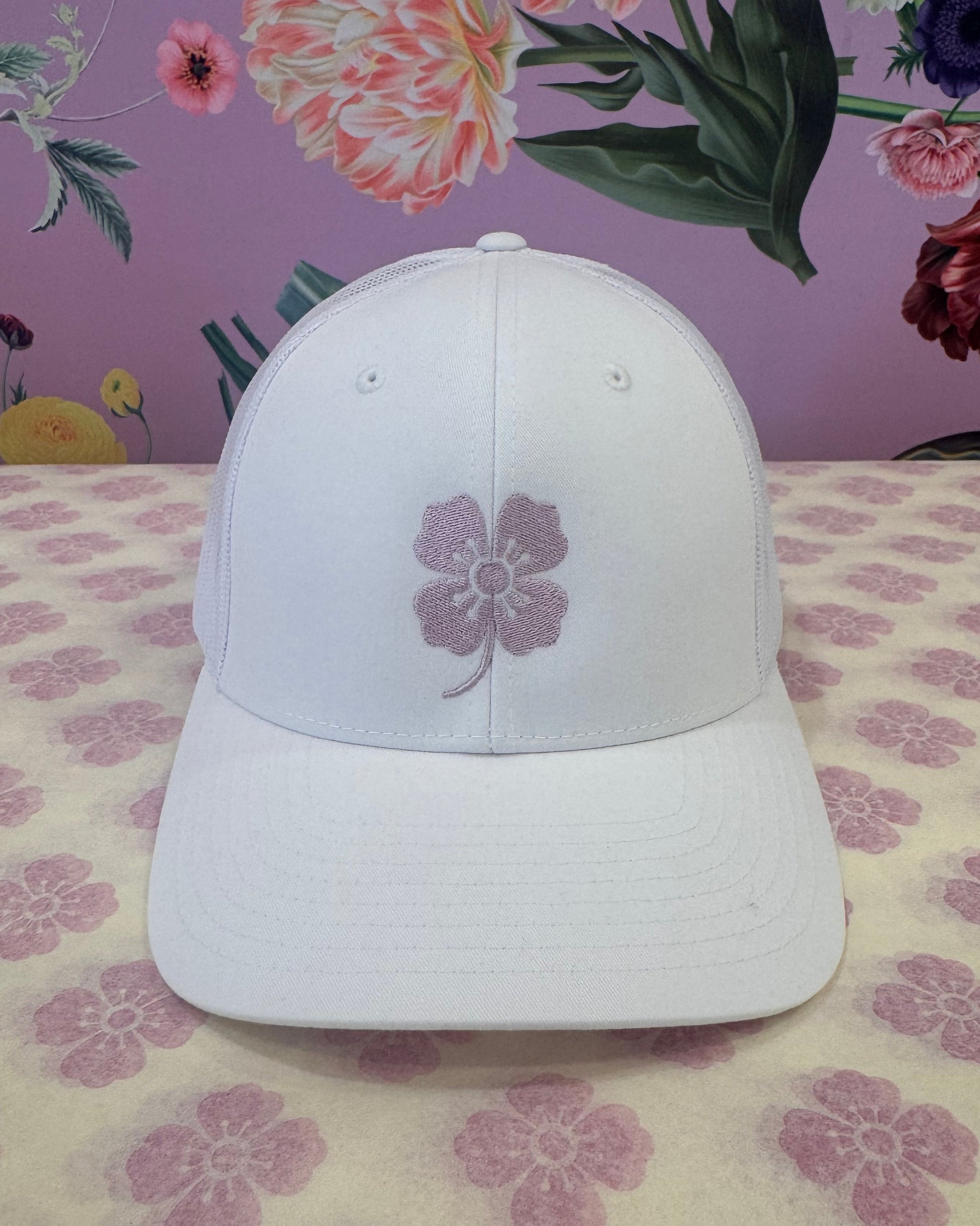 A white baseball cap with a purple embroidered flower on the front, displayed on a patterned surface with a floral wallpaper background.