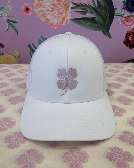 A white baseball cap with a purple embroidered flower on the front, displayed on a patterned surface with a floral wallpaper background.