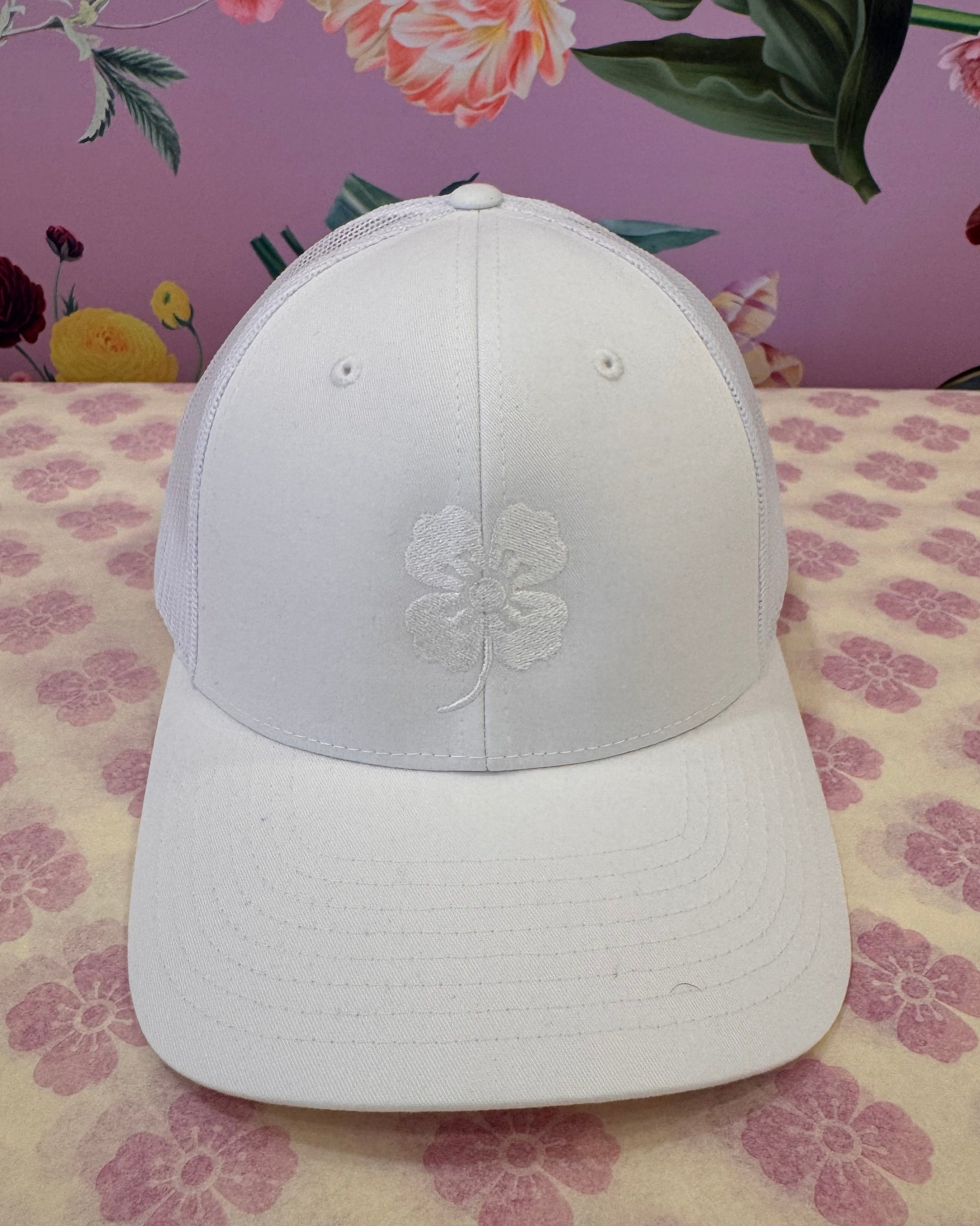 A white baseball cap with an embroidered white floral design on the front, displayed on a patterned surface with a pink floral background.