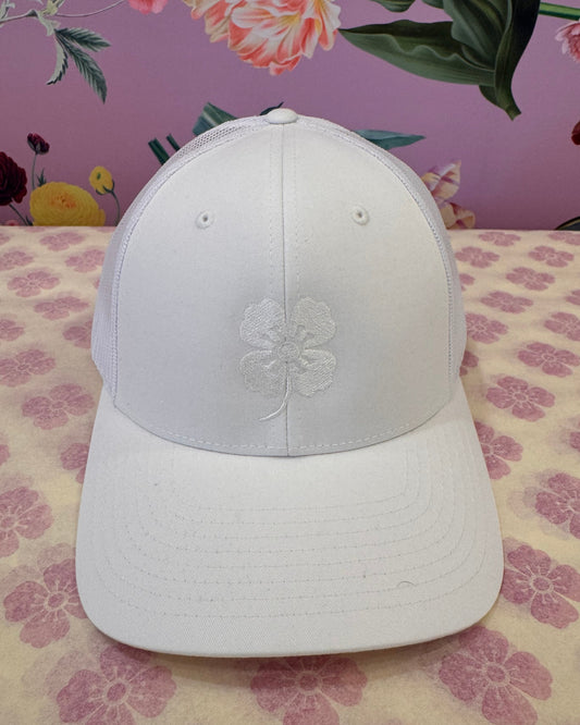 A white baseball cap with an embroidered white floral design on the front, displayed on a patterned surface with a pink floral background.